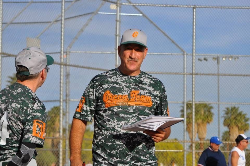 Coach holding a clipboard on a baseball field, wearing camo and orange uniform
