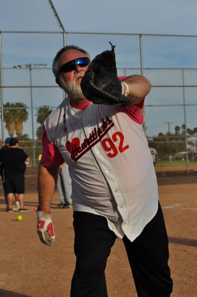 Softball player in gray and red jersey with black glove, posing on a dirt field near a chain-link fence.