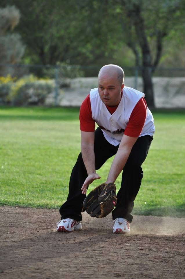 Person in red and white shirt fielding a baseball on a dirt infield, with a grassy field behind.