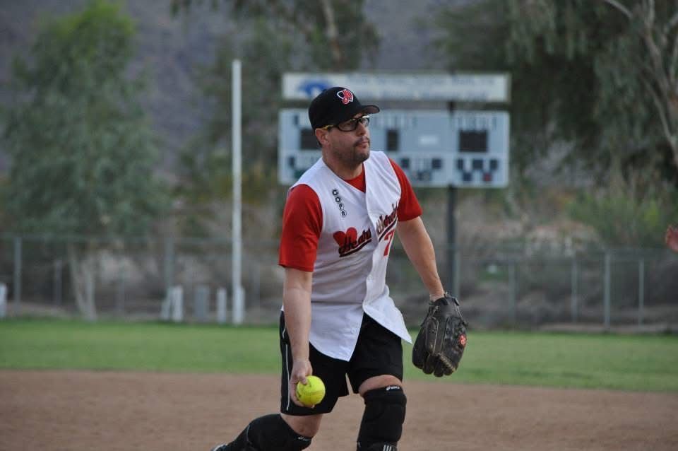 Softball player in white and red uniform winds up to throw on a dirt field