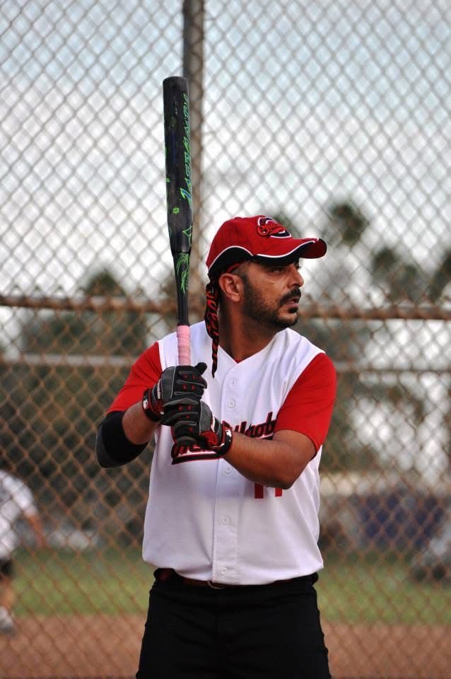 Baseball player in red and white uniform holding a bat in a batting cage, looking to the side.