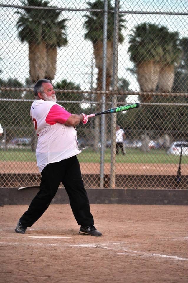 Softball batter in a white shirt and pink sleeves swinging a bat at a field with a chain-link fence and palm trees