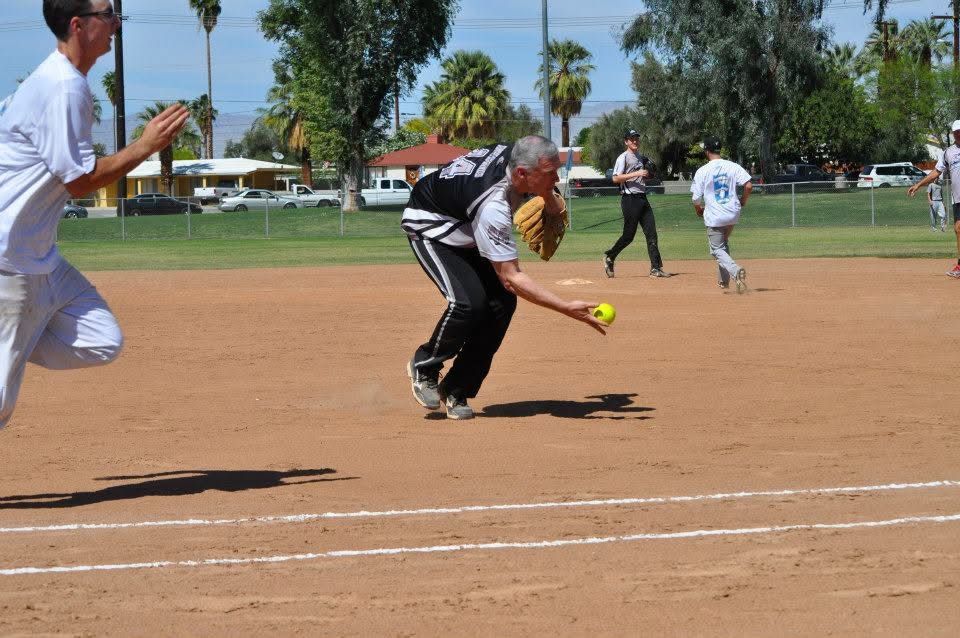 Softball player fields a rolling ball on a dirt field as teammates watch nearby.