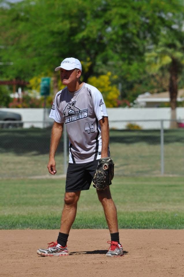 Baseball player standing on a dirt field, wearing a gray uniform, cap, and glove, with trees in the background