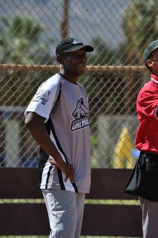 Person in a white jersey and black cap standing outdoors near a chain-link fence.