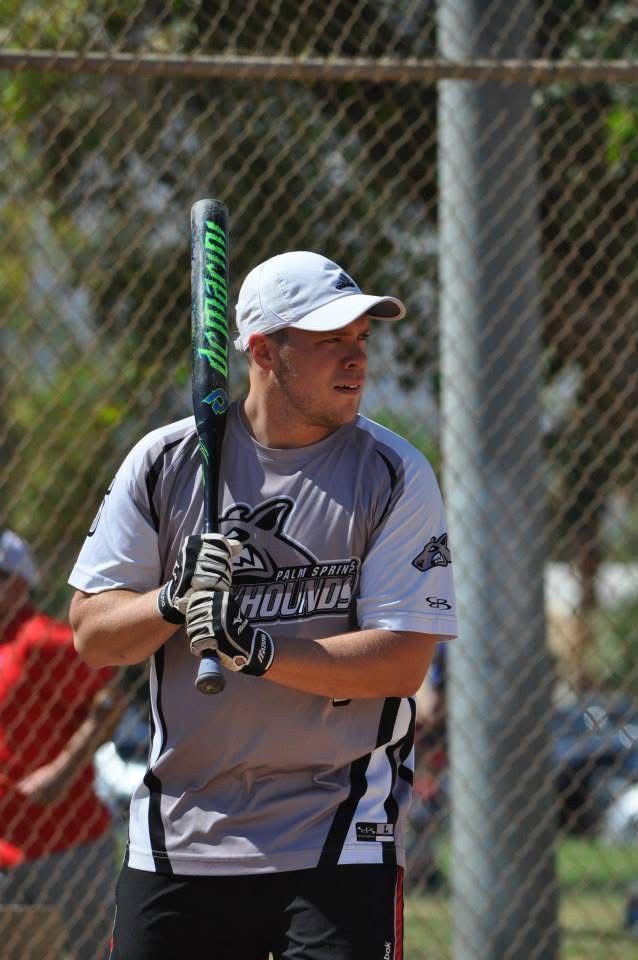 Baseball player in gray uniform holding a bat, standing near a chain-link fence on a sunny field.