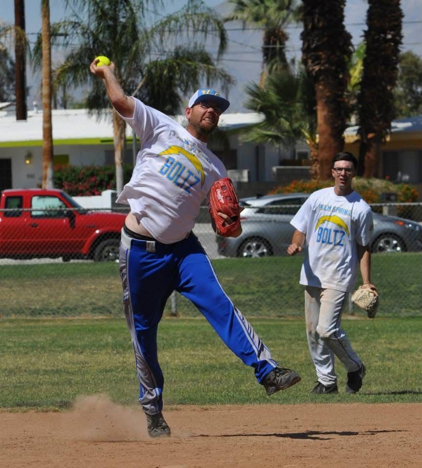 Softball pitcher in gray and blue uniform throwing a pitch on a grassy field, teammate behind him.