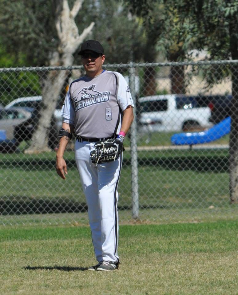 Softball player in gray uniform and black cap standing on a grassy field holding a glove.