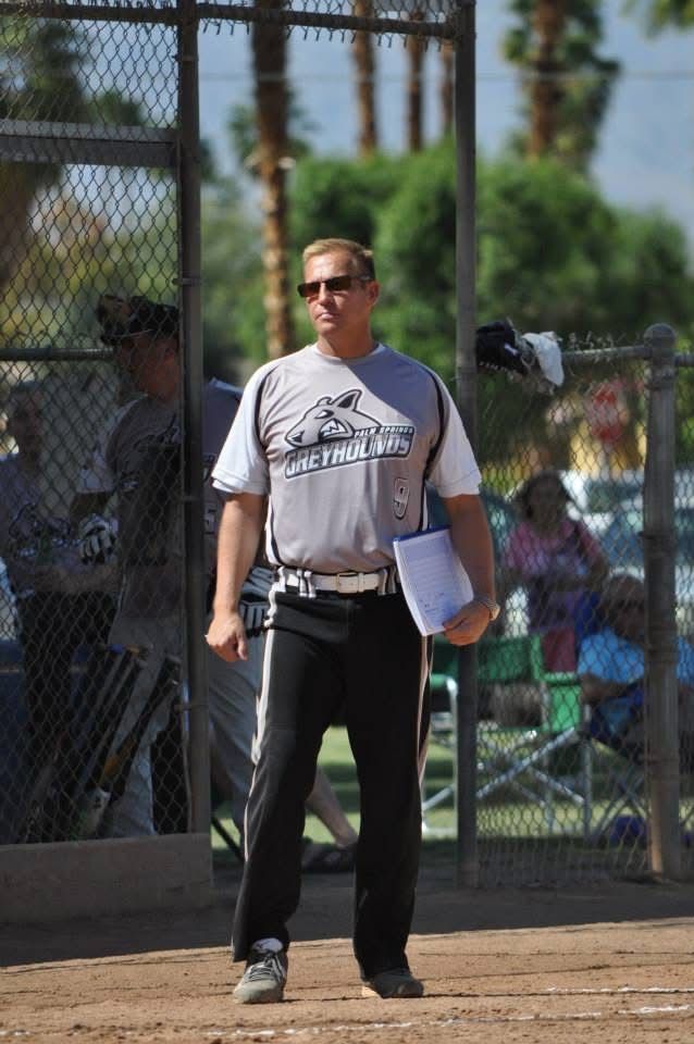 Man standing on a baseball field holding papers, with a chain-link fence and spectators in the background
