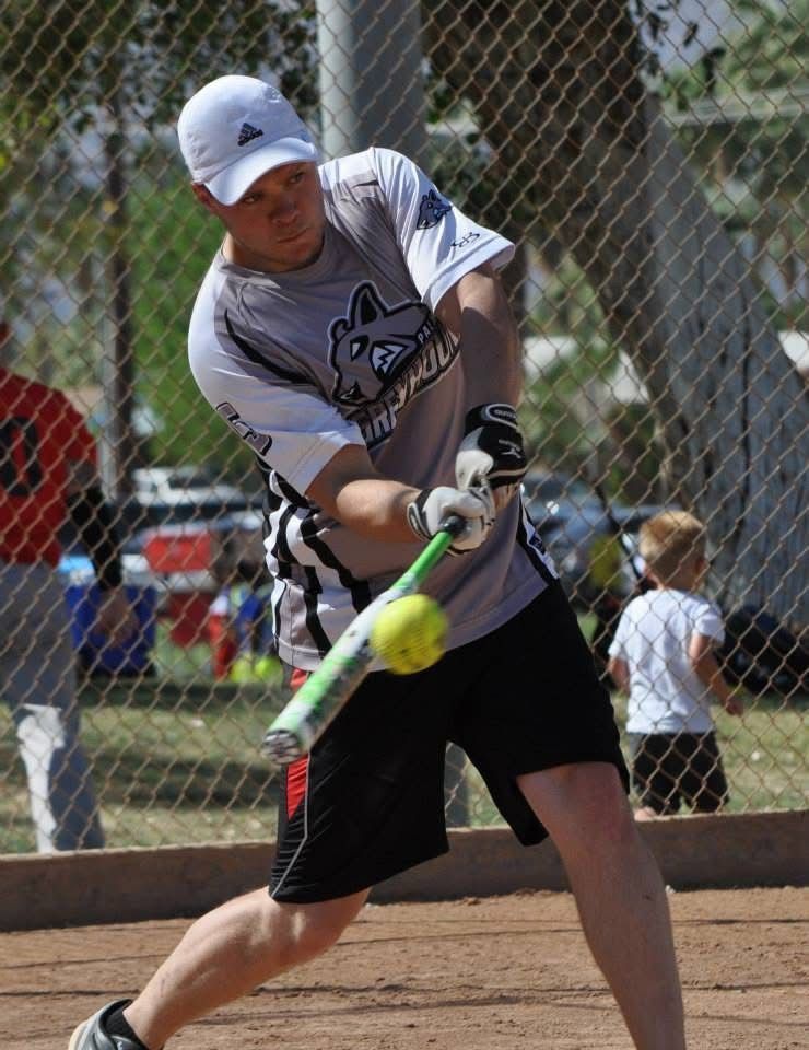 Softball player in white shirt and black shorts swinging a bat at a yellow ball on a dirt field
