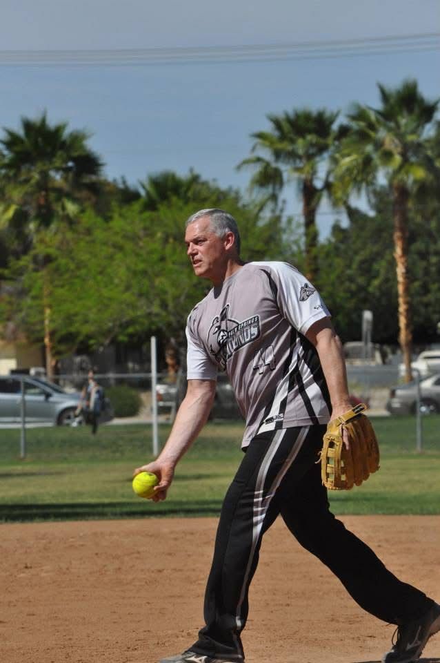Man playing softball on a dirt field, holding a yellow ball and glove with palm trees in the background
