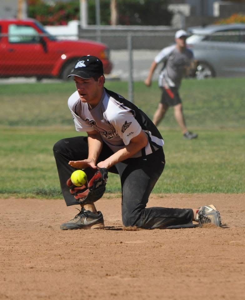 Softball player kneeling to field a yellow ball on a dirt infield, with another player in the background.