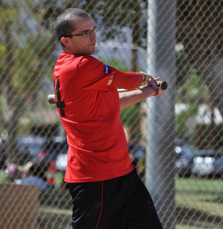 Man in a red shirt swinging a bat on a field behind a chain-link fence