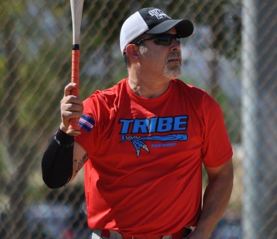 Man in red TRIBE shirt holding a baseball bat on a field, wearing a cap and sunglasses.