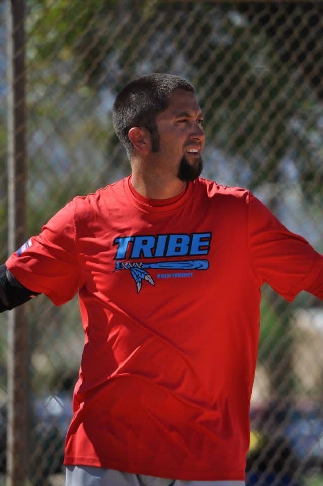 Man in a red TRIBE shirt with arms outstretched on a sports field, standing near a chain-link fence.