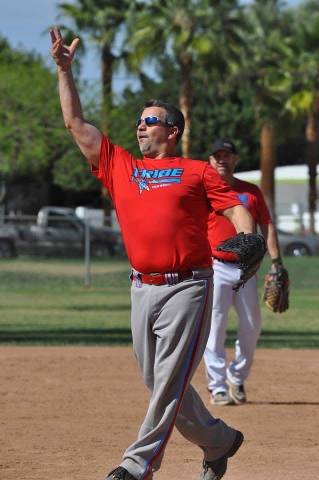 Man in red shirt and gray pants throwing a baseball on a field, with another player behind him.