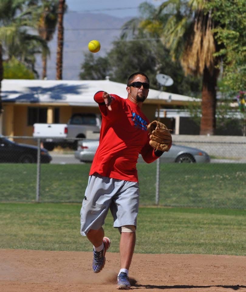 Softball player in red throws a ball on a sunny field, with a glove on the other hand.
