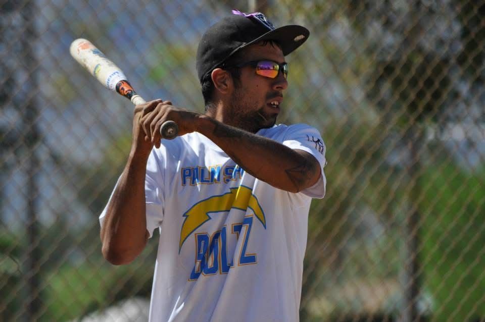 Baseball player at bat, wearing sunglasses and a white jersey, holding a bat in front of a chain-link fence