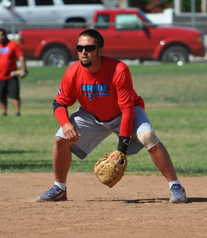 Softball infielder in red shirt crouches on dirt field, glove ready to field a ground ball