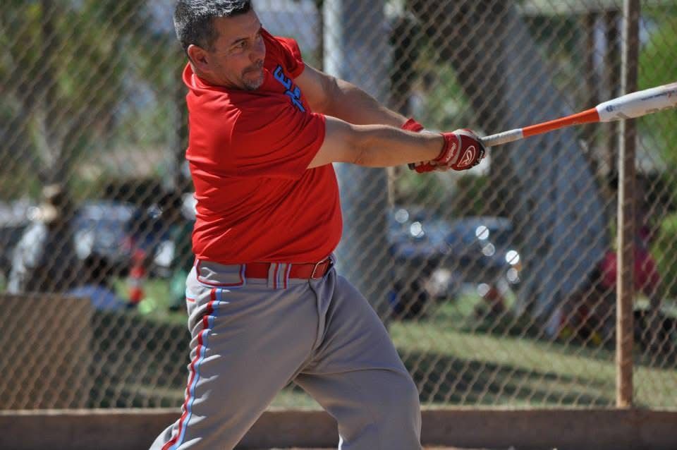 Baseball player in red shirt swinging a bat near a chain-link fence