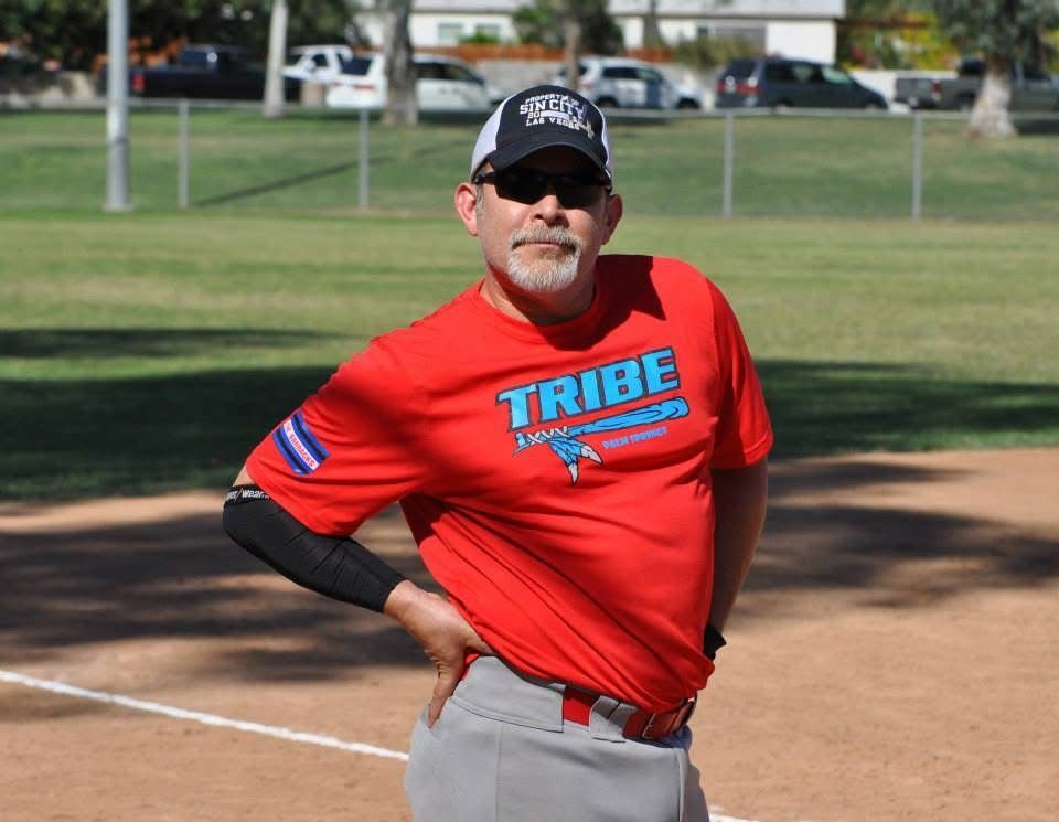 Baseball coach in a red TRIBE shirt and black cap stands hands on hips on a field, with a glove in his back pocket