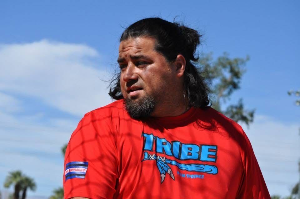 A man in a red shirt stands outdoors under a blue sky, looking to the side.