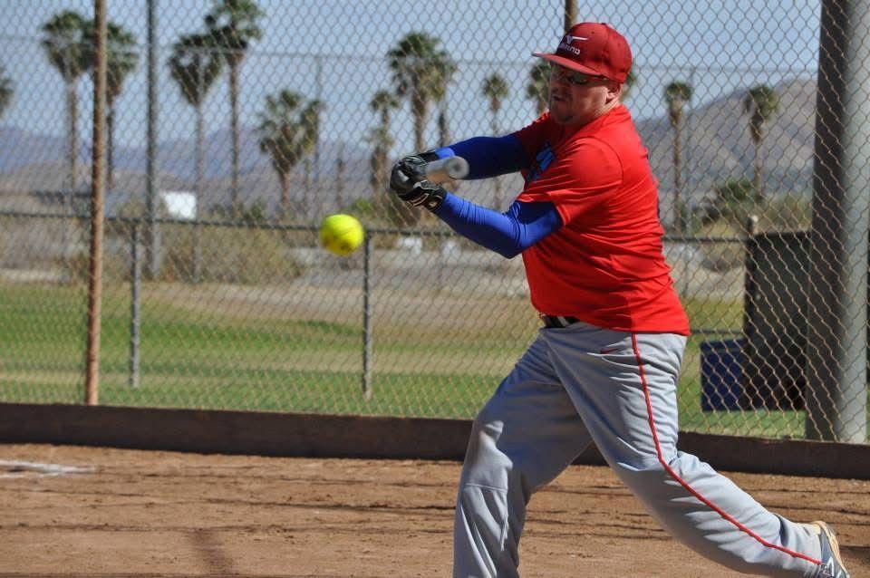 Baseball player in red swings at a pitch on a dirt field near a chain-link fence.