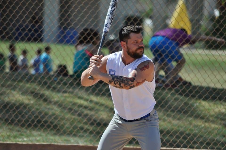 Baseball player in white jersey swings a bat on a field with spectators behind a fence