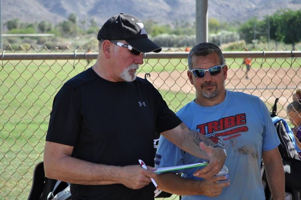 Two men outdoors by a chain-link fence, one holding a clipboard and gesturing while the other listens.