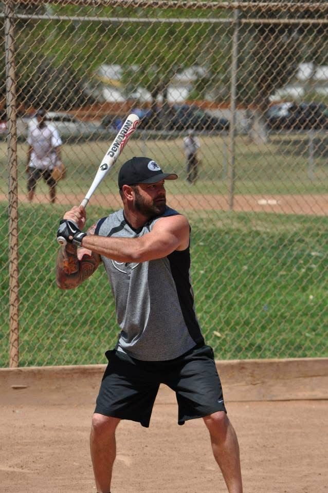 Baseball batter at the plate holding a bat, with a chain-link fence and field in the background