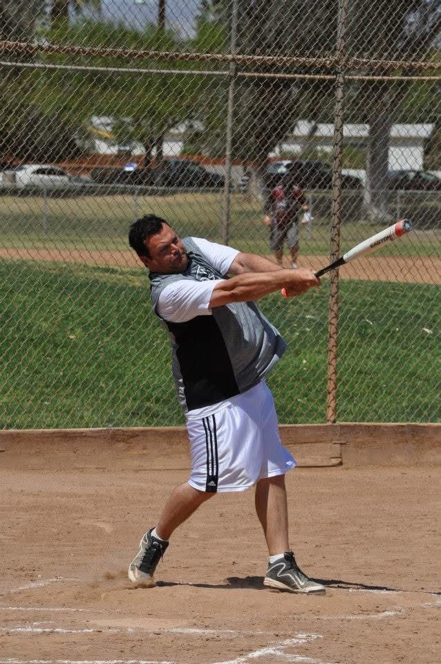 Baseball player swinging a bat on a dirt field, with a chain-link fence and grass in the background.