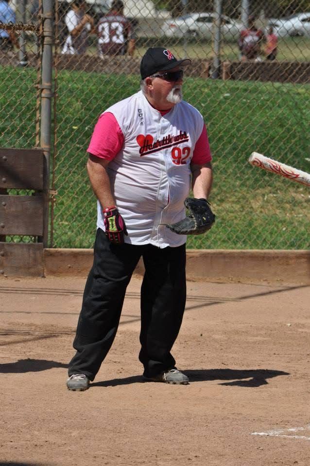 Softball player in white and pink jersey stands on a dirt field holding a glove near a chain-link fence.