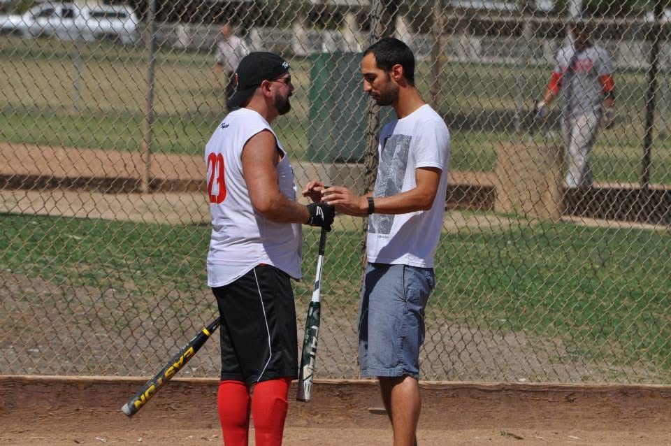 Two softball players in white jerseys talk beside a chain-link fence on a dirt field.