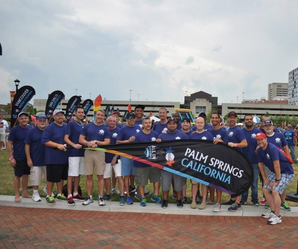 Group of runners in blue shirts holding a “Palm Springs, California” banner at a race finish line.