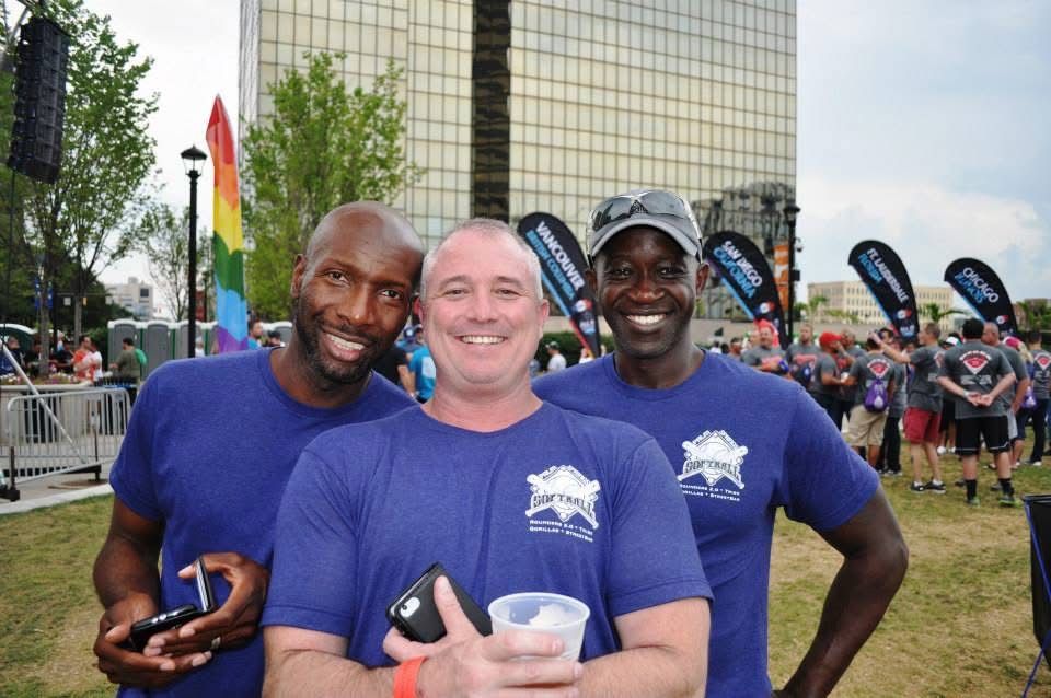 Three smiling runners in blue shirts posing at a race finish area with crowd and flags behind them