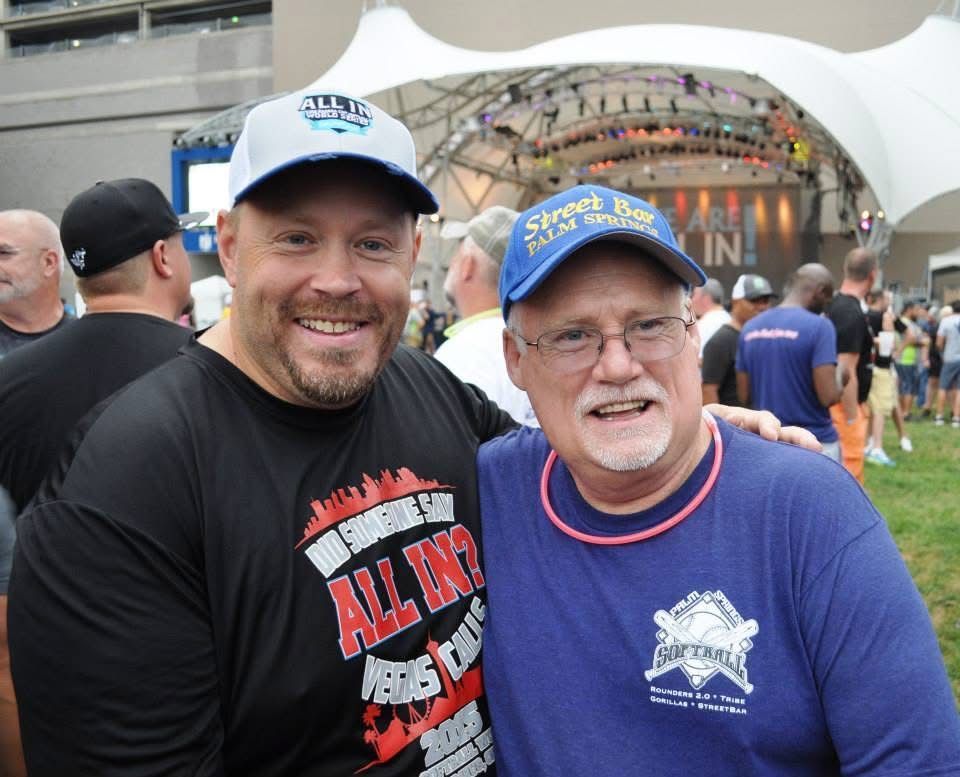 Two smiling men in caps and event shirts posing together outdoors at a crowded festival stage