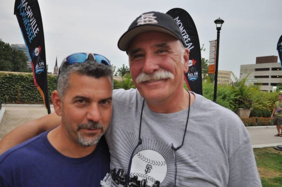 Two smiling men pose outdoors, one wearing a black cap and the other blue sunglasses on his head.