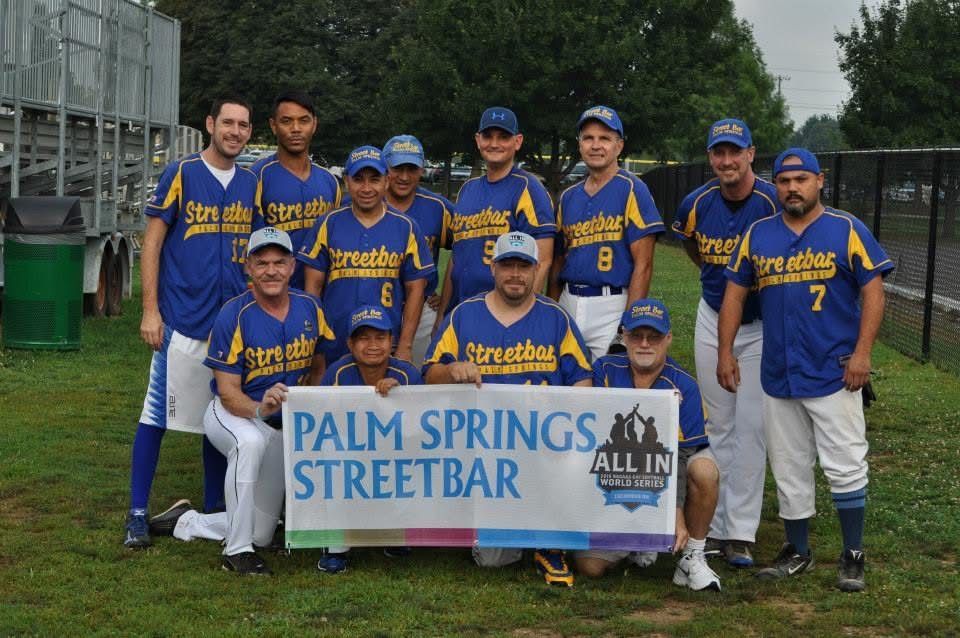 Softball team in blue uniforms posing on grass behind a Palm Springs Streetbar sponsor banner.