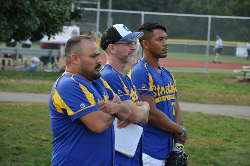 Three men in blue and yellow sports uniforms standing with arms crossed at a baseball field