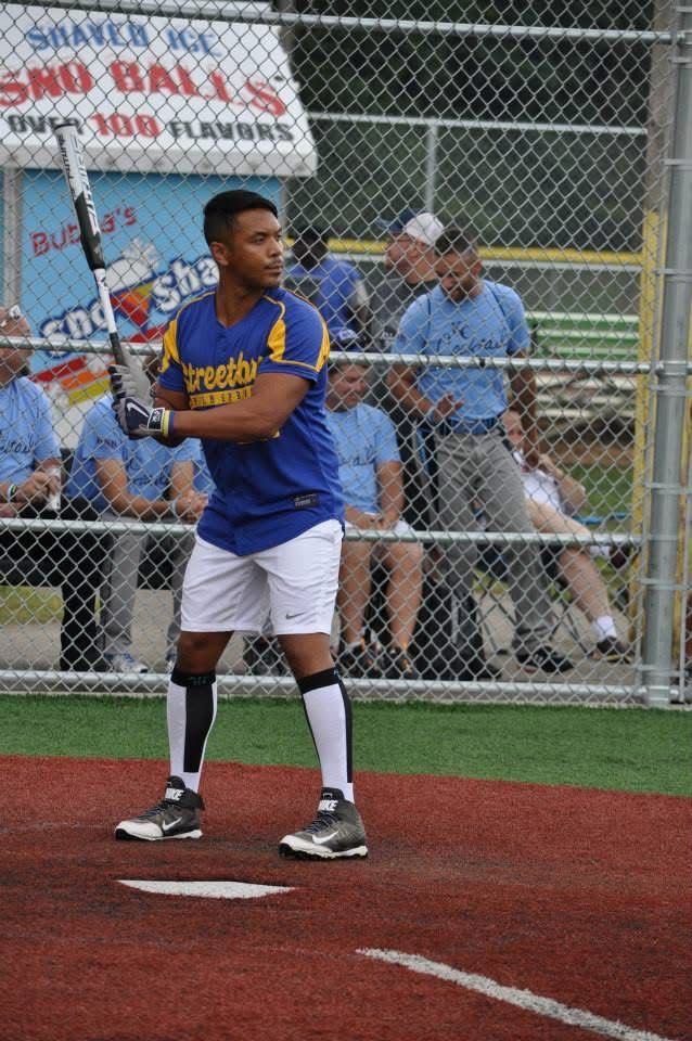 Softball batter in blue uniform stands in the batter’s box, ready to swing near the fence.