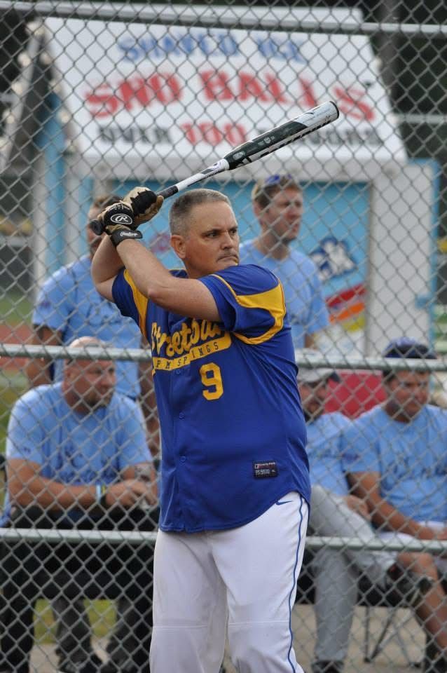 Softball player in blue and yellow jersey swings bat at home plate behind chain-link fence
