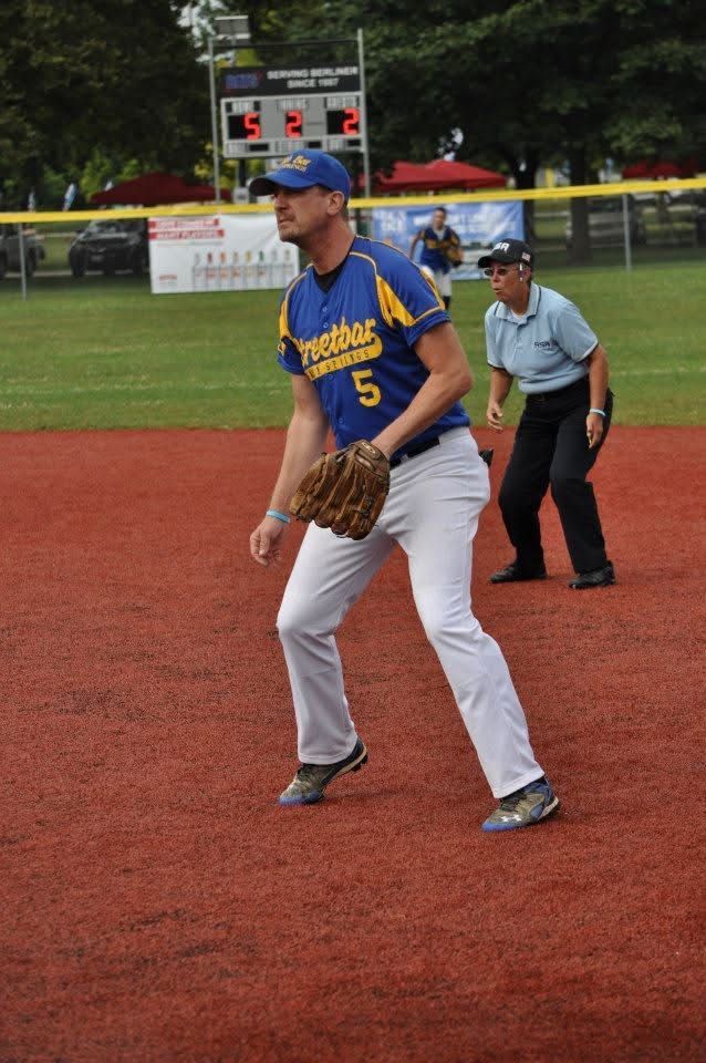 Baseball player in blue jersey and gray pants fielding on a red infield, with an umpire behind him.