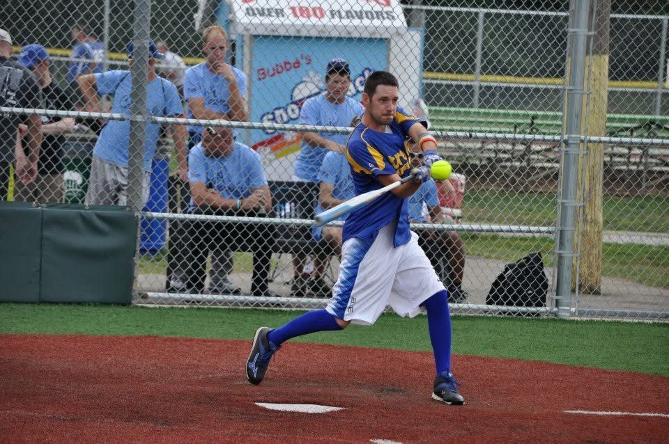 Softball player in blue uniform swings at a yellow ball on a red field, with spectators behind a chain-link fence