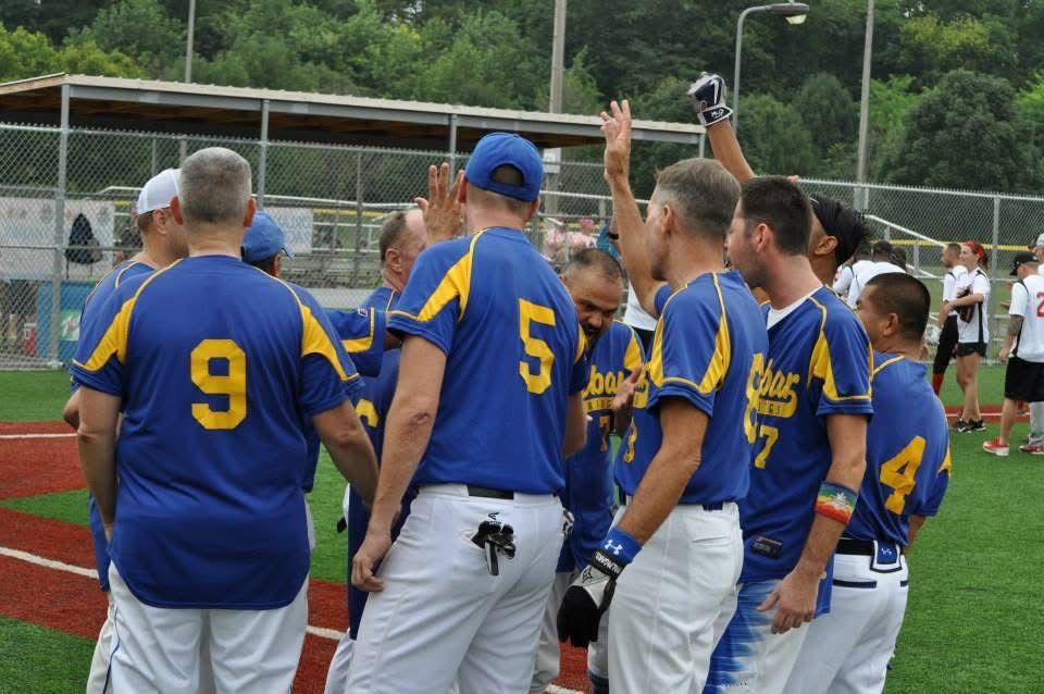 Baseball team in blue jerseys huddles on a field, one player raising a hand near the dugout.