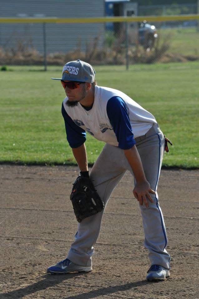Baseball player in gray and blue uniform crouches in ready stance on a dirt field.