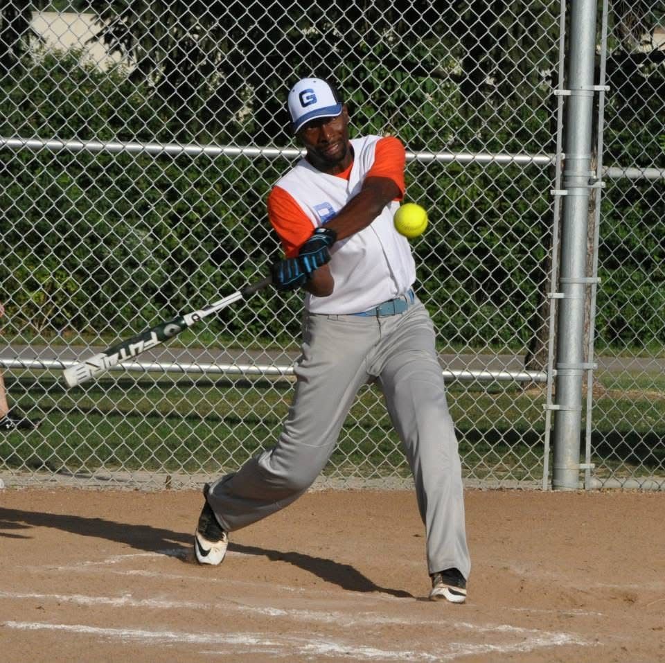 Softball player in gray and orange swings at a yellow ball on a dirt field near a chain-link fence