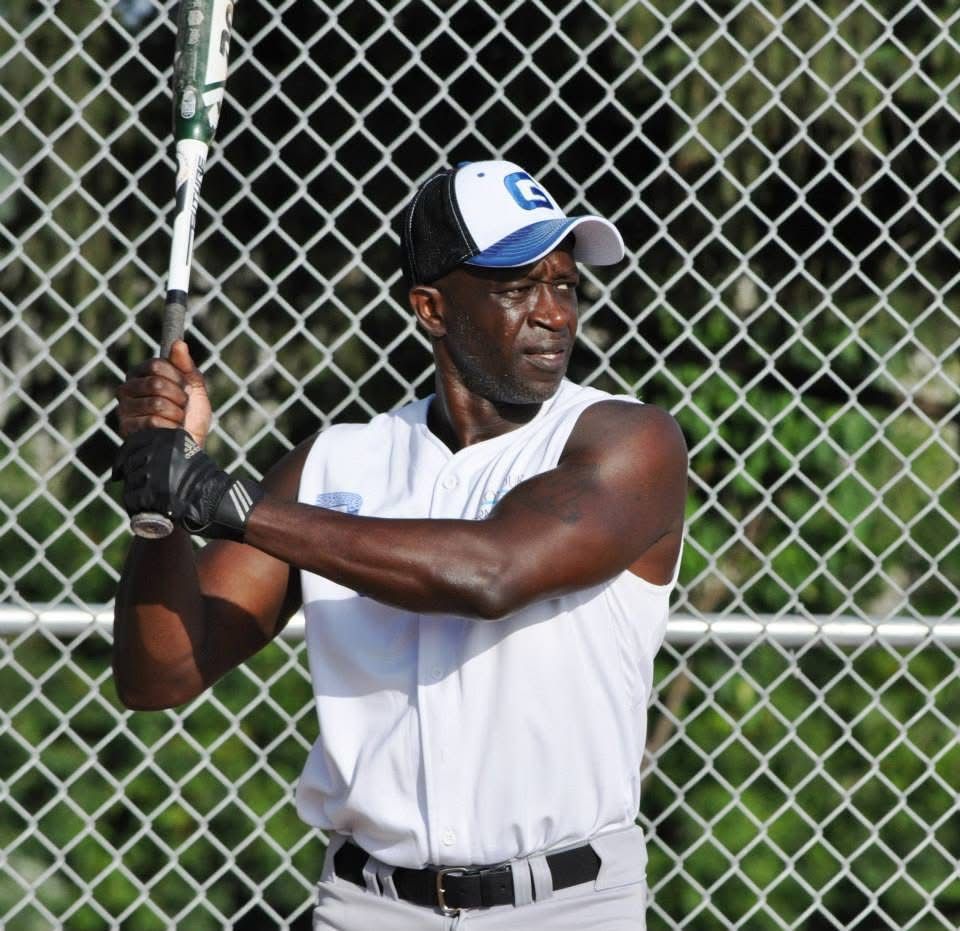 Man in white sleeveless shirt holding a baseball bat at a batting cage, preparing to swing
