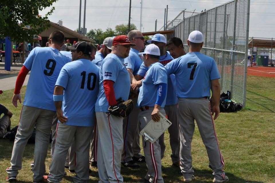 Baseball team huddled on grass near a chain-link fence at a field, wearing light blue jerseys and gray pants
