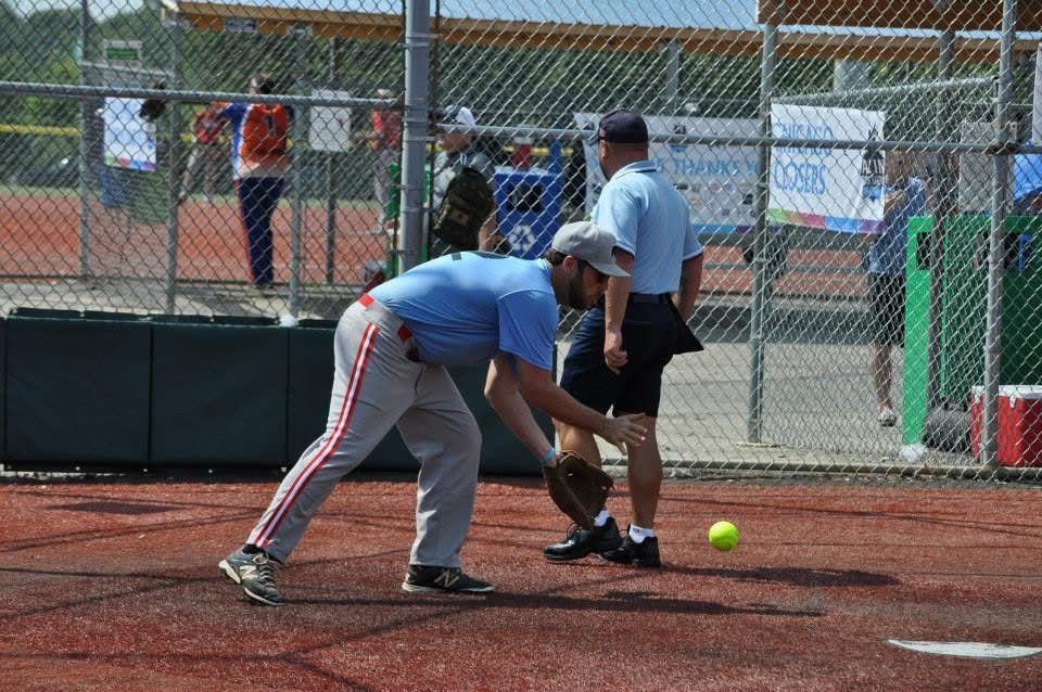 Two players crouch over a softball on a field, with a coach nearby and a chain-link fence behind them