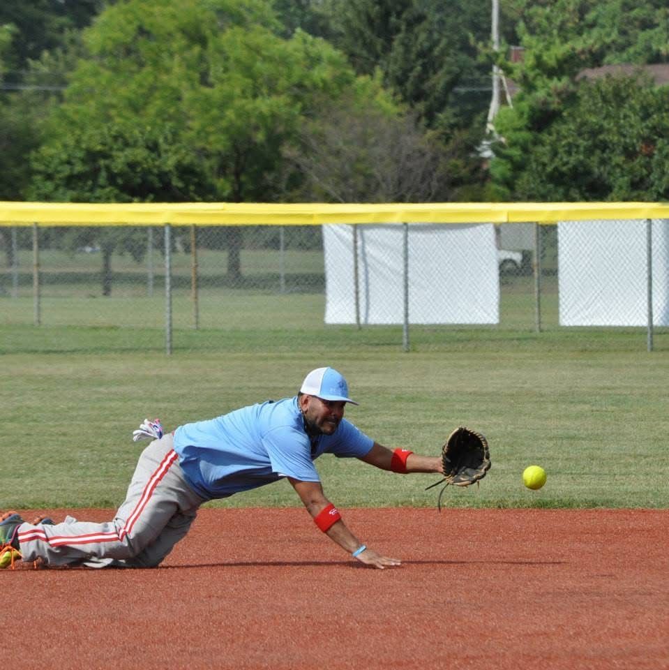 Softball player diving toward a ball on a dirt field, glove outstretched near the base path.